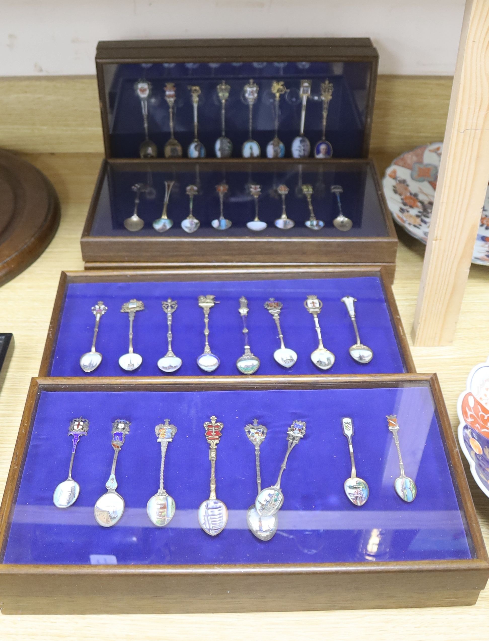 A collection of sixty four 800 standard silver (many gilt) and enamel souvenir spoons, early 20th century, in eight display cases, Including views of London, Germany, Italy, Austria, Spain etc.